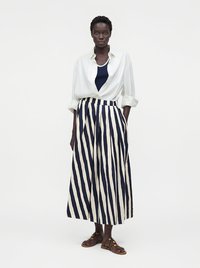 Femme portant une chemise blanche boutonnée sur un top bleu marine, un pantalon large rayé vertical bleu marine et blanc, et des sandales marron, debout avec une main dans la poche.