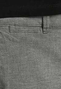 Tissu gris texturé du pantalon, présentant une surface lisse avec de petits motifs diagonaux. Comprend une ceinture standard et des passants pour ceinture.