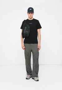 Zwarte T-shirt met blauwe tekst, grijze cargobroek met grote zakken, zwarte schoudertas met rits, zwarte pet en grijze sneakers.