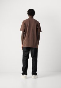 Man with short dreadlocks wearing a brown T-shirt, black pants, and white sneakers, standing facing a plain white background.