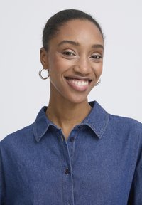 Chemise en denim bleu foncé avec col classique, devant boutonné et surpiqûres contrastantes. Le modèle porte de petites boucles d'oreilles créoles.