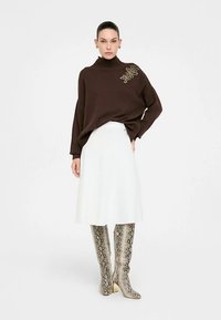 Brauner, gerippter Strickpullover mit hohem Kragen und Verzierungen, kombiniert mit einem weißen ausgestellten Rock und kniehohen Stiefeln mit Schlangenmuster.