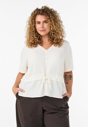 Žena s kučeravými vlasmi v textúrovanom bielom zapínanom top s krátkymi rukávmi a tmavých nohaviciach, stojaca s rukami v vreckách a ukazujúca tetovanie ruže.