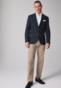 Blazer de un azul marino oscuro, camisa blanca, pantalones beiges, zapatos negros. El blazer tiene dos botones y bolsillos; la camisa tiene un cuello doblado y puños.