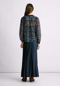 Blusa trasparente a maniche lunghe di colore blu navy con motivo floreale e yoke nero solido, abbinata a una gonna maxi in denim scuro e stivali con tacco marroni.