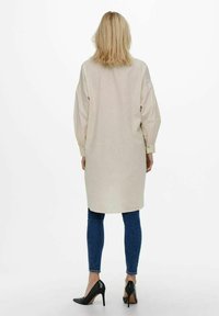 Beige Hemdblusenkleid mit langen Ärmeln in lockerer Passform und elastischen Bündchen, kombiniert mit dunkelblauen Skinny Jeans und schwarzen High Heels.