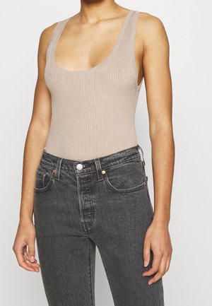 Osoba nosící béžové žebrované tílko zastrčené do černých džín s vysokým pasem, stojící před jednoduchým pozadím.