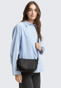Borsa a tracolla nera con texture, dalla forma curva e dettagli con frange, indossata con una camicia a righe blu e bianche e pantaloni scuri.