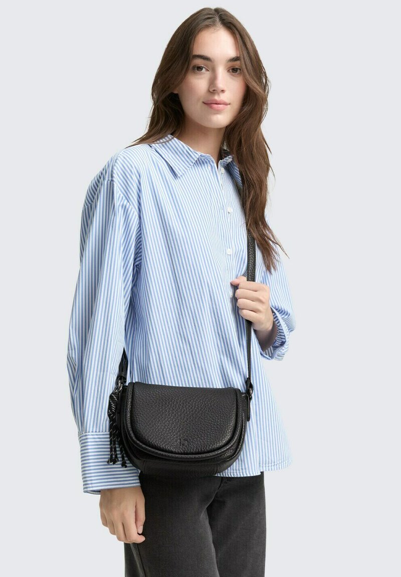 Borsa a tracolla nera con texture, dalla forma curva e dettagli con frange, indossata con una camicia a righe blu e bianche e pantaloni scuri.