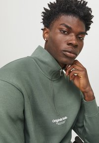 Sudadera verde con cremallera y cuello alto, que presenta un logo blanco en la parte delantera, fabricada en un tejido suave y texturizado.