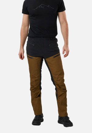 Uomo che indossa pantaloni da trekking neri e marroni con tasche con cerniera, t-shirt nera con motivo montagna e scarpe da trekking nere, in piedi.