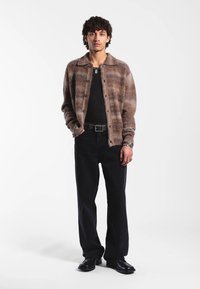 Ruskea ruudullinen paita takki kauluksella, päällä mustan paidan kanssa, yhdistettynä mustiin leveälahkeisiin housuihin ja mustiin kenkiin. Mukana hopeiset asusteet.