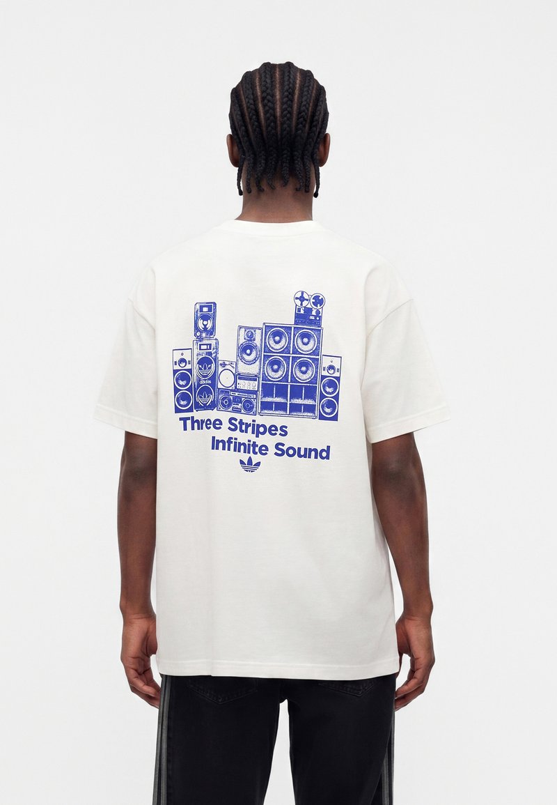 Homme aux cheveux tressés portant un t-shirt blanc avec une illustration bleue représentant une pile d'enceintes et le texte « Three Stripes Infinite Sound » au dos, debout devant un fond blanc.
