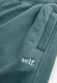 Groene katoenen joggingbroek met elastische tailleband, voorzien van een zijzak en witte geborduurde tekst "self." nabij de zoom. Soepele textuur.