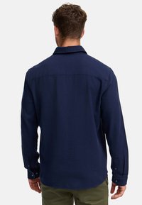 Camisa de manga larga azul marino hecha de un tejido texturizado, con cuello puntiagudo y puños de botón, vista desde la parte posterior.