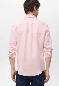 Lichtroze blouse met lange mouwen, met een button-down kraag, plooien aan de achterkant en opgerolde manchetten, gemaakt van ademende stof. Gedorven met donkerblauwe jeans.
