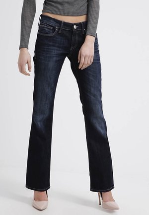 Kobieta nosząca ciemnoniebieskie spodnie typu bootcut, szary prążkowany długi rękaw crop top oraz beżowe buty na wysokim obcasie, stojąca na jednolitym tle.