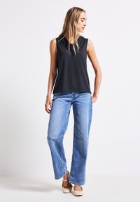 Top nero senza maniche con un fit rilassato, abbinato a jeans larghi di colore blu chiaro. Le calzature sono piatte e di colore crema, con cinturini alla caviglia.