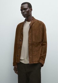 Veste en daim brun avec un design à bouton, dotée d'un petit col et d'une poche latérale, superposée à un pull beige et un pantalon sombre.