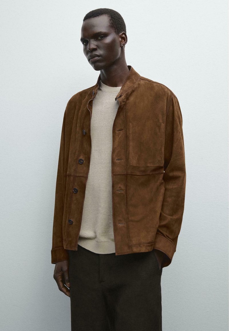 Veste en daim brun avec un design à bouton, dotée d'un petit col et d'une poche latérale, superposée à un pull beige et un pantalon sombre.