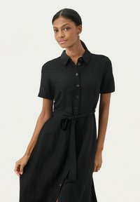 Abito camicia in lino nero con maniche corte, chiusura frontale a bottoni e vita con cintura. Presenta una leggera fessura e una texture liscia. Design semplice e classico.