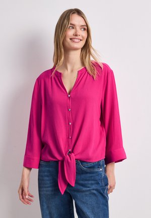 Frau mit langen blonden Haaren, die eine knallrosa geknöpfte Bluse trägt, die an der Taille gebunden ist, blaue Jeans, lächelnd und nach rechts schauend.