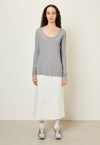 Mlada ženska s kodrastimi lasmi, oblečena v sivo majico z dolgimi rokavi, belo midi krilo, bele nogavice in sive superge, ki stoji ob svetlem zidu.