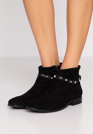 Botines de ante negro con tiras tachonadas de plata, usados sobre piernas desnudas sobre un fondo claro.
