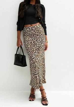 Vrouw draagt een midi rok met luipaardprint, een zwart cropped top, een zwarte cardigan, zwarte sandalen met bandjes en houdt een zwart handtasje vast tegen een witte achtergrond.