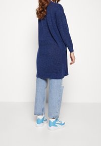 Lång, mörkblå cardigan med ribbade muddar och fåll, kombinerad med ljusblå slitna jeans och vita sneakers med blå detaljer.