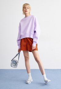 Lavender cropped sweatshirt with Nike logo, orange shorts, white socks, and silver sneakers. Also features a small grey crossbody bag.