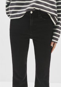 Osoba v černých rovně střižených džínech a dlouhém šedém a bílém pruhovaném tričku, ruka položená podél těla.