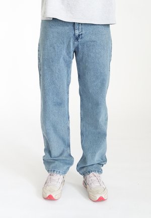 Jean droit bleu clair en denim, doté d'une taille visible et de détails de couture subtils, associé à des chaussures de sport blanches.