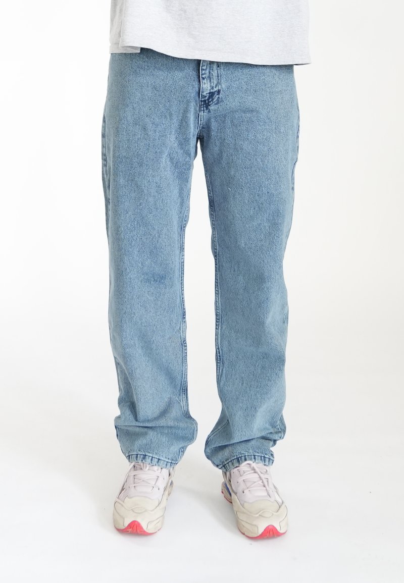 Jean droit bleu clair en denim, doté d'une taille visible et de détails de couture subtils, associé à des chaussures de sport blanches.