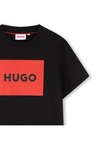 Svart T-shirt i bomull med rund halsringning. Har en framträdande röd rektangel med texten "HUGO" i fet svart stil. Kortärmad.