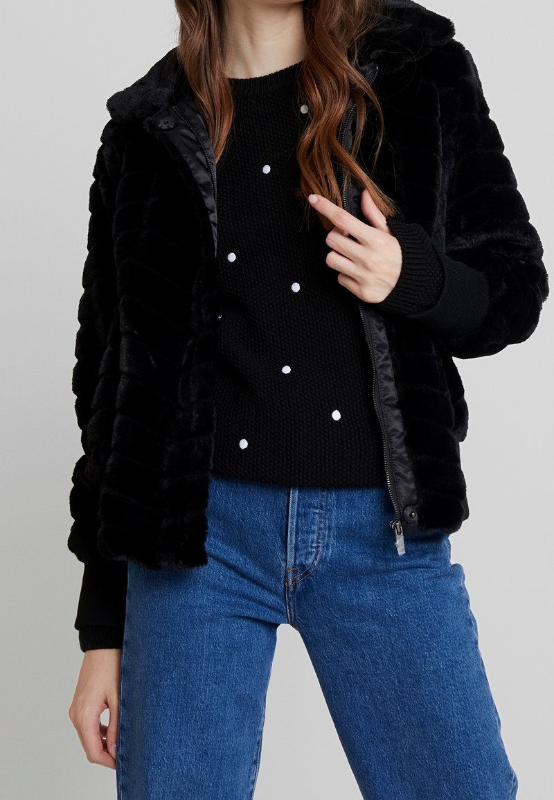 Femme portant une veste en fausse fourrure noire sur un pull noir à pois blancs et un jean bleu, main ajustant le zip de la veste.