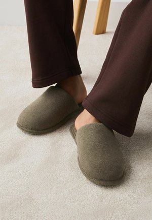 Inimene, kes kannab pruune pükse, libistab jalad hallidesse, fliisvoodriga, kinniste varvastega sussidesse helel vaibal, puidust toolijalgade kõrval.