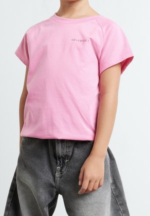 Enfant portant un t-shirt rose clair à manches courtes avec le logo "Converse" et un jean ample gris foncé, debout devant un fond uni.
