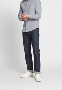 Vaaleansininen pitkähihainen paita, jossa napillinen kaulus ja rintatasku. Tummat suorat farkut, lahkeet kääräisty. Valkoiset tennarit kuvioiduilla pohjilla.