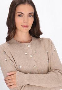 Beige stickad kofta med ribbad textur, prydd med pärl- och ädelstensdetaljer, samt rund halsringning med knappstängning.