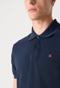 Nærbillede af en person iført en mørkeblå poloshirt med to knapper og et lille lyserødt „cK“-logo på brystet.