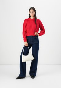 Röd stickad tröja, högmidjade mörkblå jeans, vit axelväska med snörningsdetalj och svarta skor. Stående mot en enfärgad bakgrund.