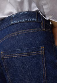 Mörkblå denimjeans har kontrasterande orange sömmar, en platt bakficka och en etikett med "ADOLFO DOMINGUEZ" nära midjan.