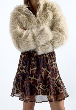 Personne portant une veste en fausse fourrure beige avec une jupe à imprimé léopard, bras croisés, sur un fond blanc uni.