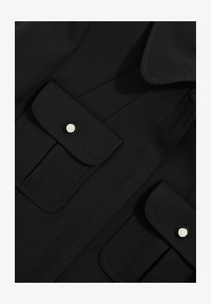 Svart tygskjorta med två bröstfickor med tryckknappar, strukturerad krage och texturerad yta. Rena linjer och minimal design.