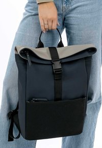 Zaino roll-top blu navy e grigio realizzato in tessuto con una texture opaca, dotato di una tasca frontale e spallacci regolabili.