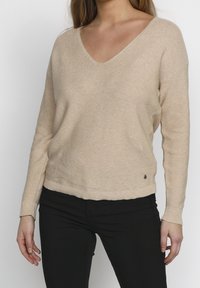 Beige stickad tröja med V-ringning, långa ärmar och ribbade muddarna. Har en liten logotyp vid nederkanten och en avslappnad passform.