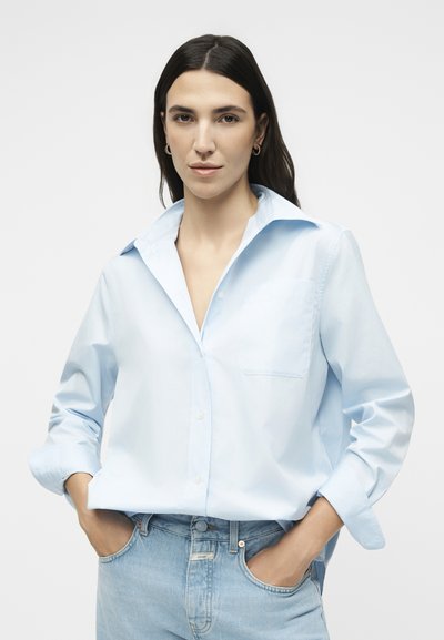 Γυναίκα με μακριά σκούρα μαλλιά που φορά χρυσούς κρίκους, ανοιχτό μπλε πουκάμισο με κουμπιά και γυρισμένα μανίκια και ανοιχτό μπλε τζιν, τα χέρια στις τσέπες.