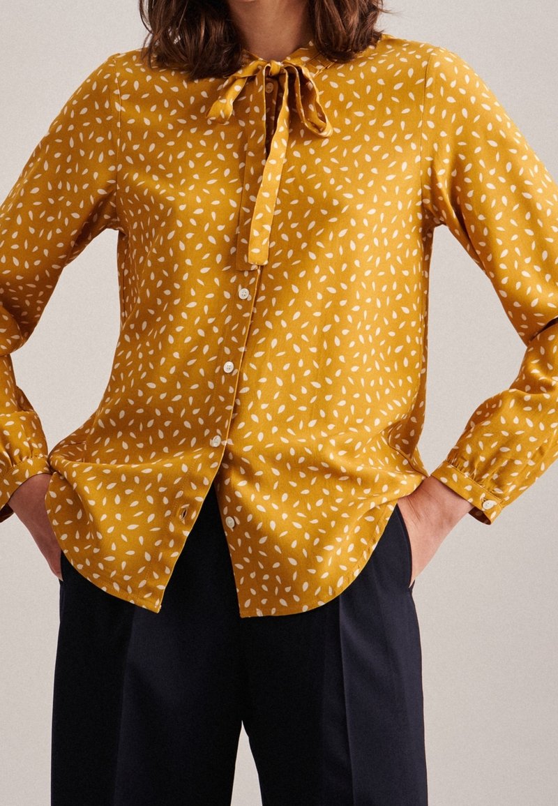 Yellow blouse with white leaf print, features a tie collar and button-down front, long sleeves, and slightly rounded hem. Textured fabric.