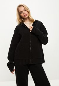 Černá zipová mikina vyrobená z jemné látky, s kapucí, žebrovanými manžetami a dvěma předními kapsami se strukturovaným lemem.